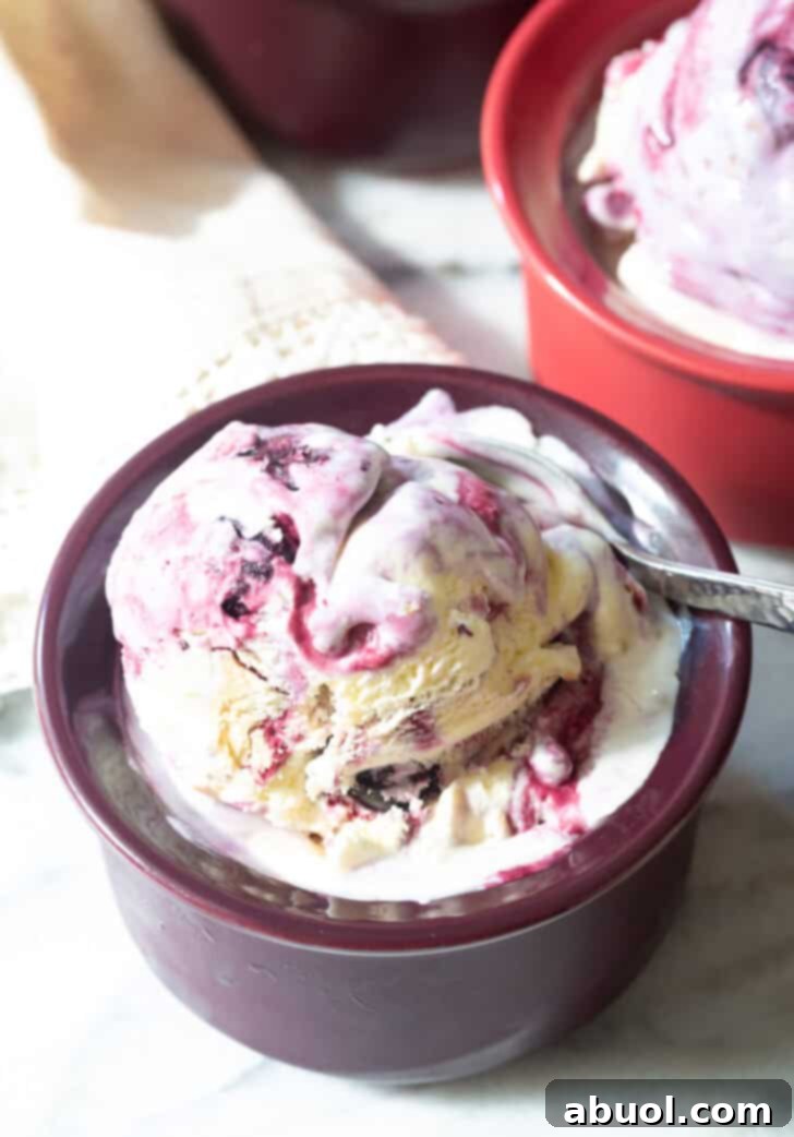 Dark bowl with blueberry pie ice cream. A spoon is taking a scoop from the bowl, showing the swirling blueberry and pie crust pieces.