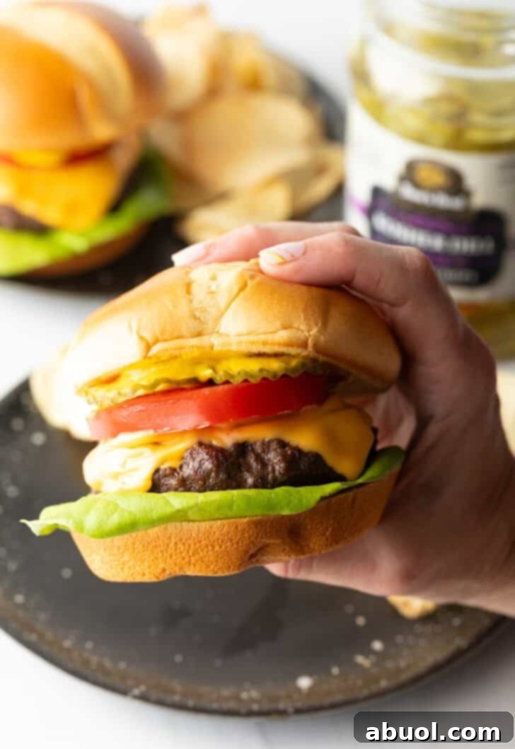 Air Fryer Burger Perfection 2 Hand holding a juicy air fryer hamburger loaded with cheese, lettuce, tomatoes and pickle slices.