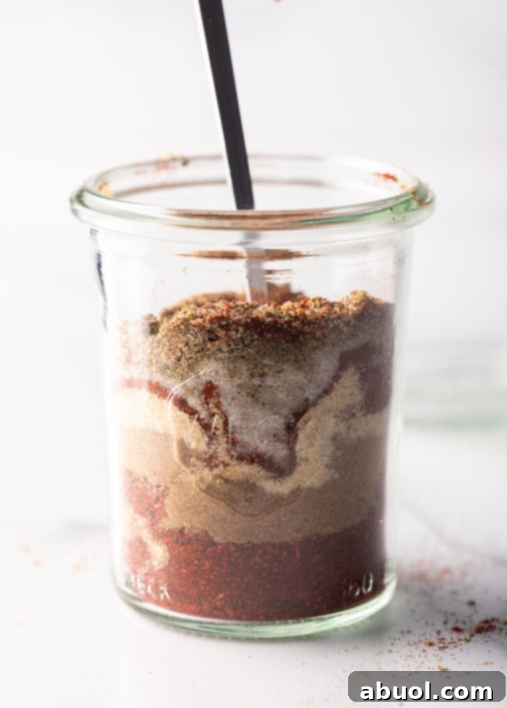 A spoon actively stirring the layers of chicken taco spices in a tall glass jar, illustrating the mixing process.
