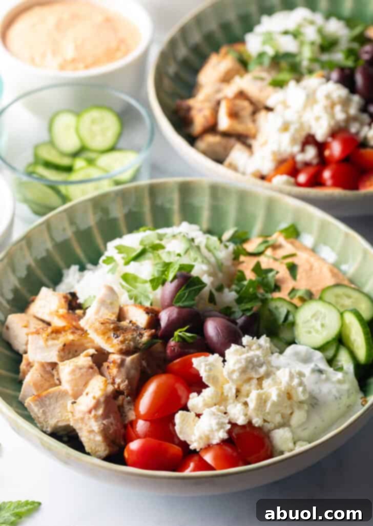 A beautifully arranged Greek chicken bowl featuring grilled chicken, white rice, halved cherry tomatoes, sliced cucumbers, Kalamata olives, a dollop of tzatziki, and crumbled feta cheese.