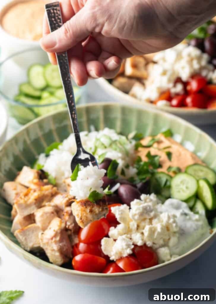 A fork scooping a generous bite from a Greek chicken bowl, highlighting the tender chicken, rice, and fresh vegetables.