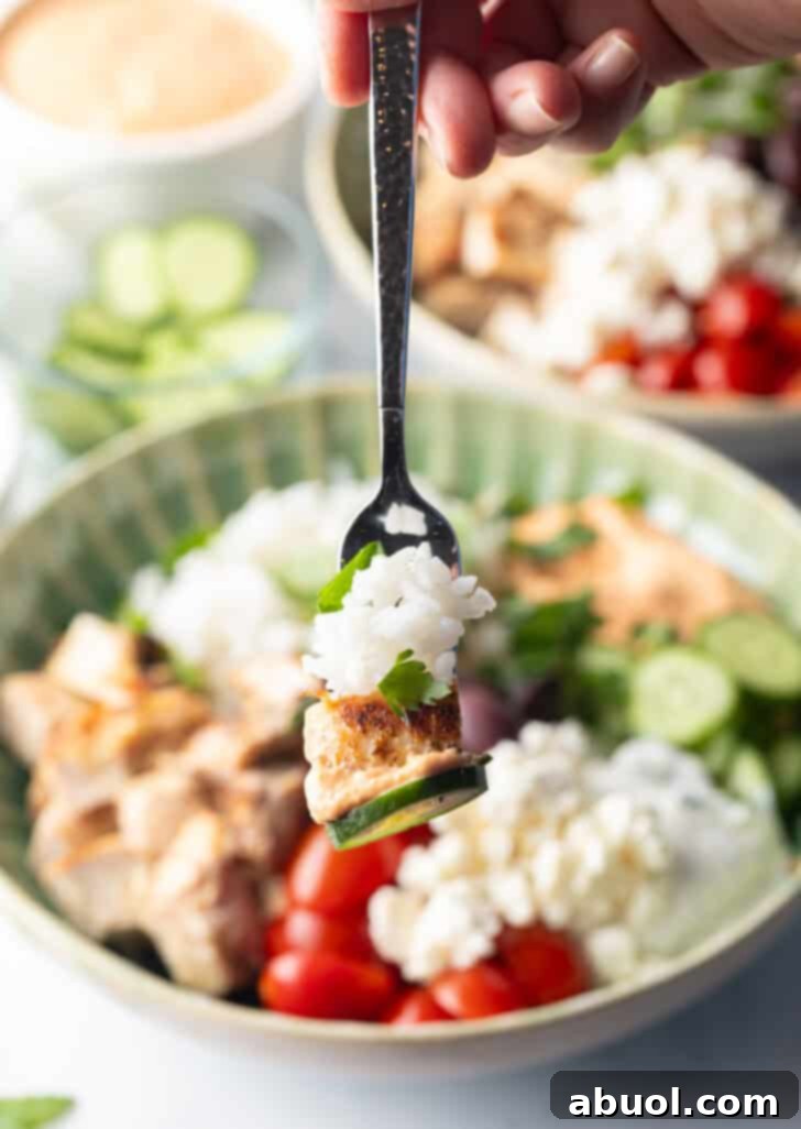 A fork holding a bite of rice and cucumber, lifted from a colorful Greek chicken bowl.