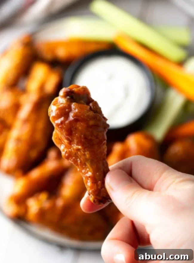 A hand holds up a perfectly baked buffalo chicken wing drumette, showcasing its crispy skin and rich sauce, enticing viewers to grab one.