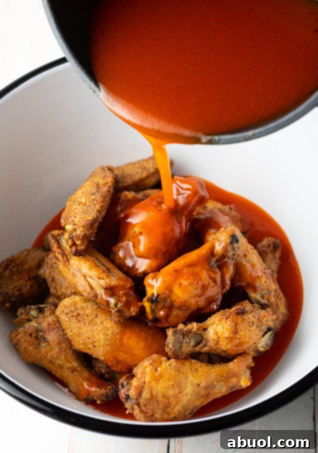 A pristine white mixing bowl containing golden-brown, baked chicken wings, with a rich, vibrant buffalo sauce being poured generously over them.