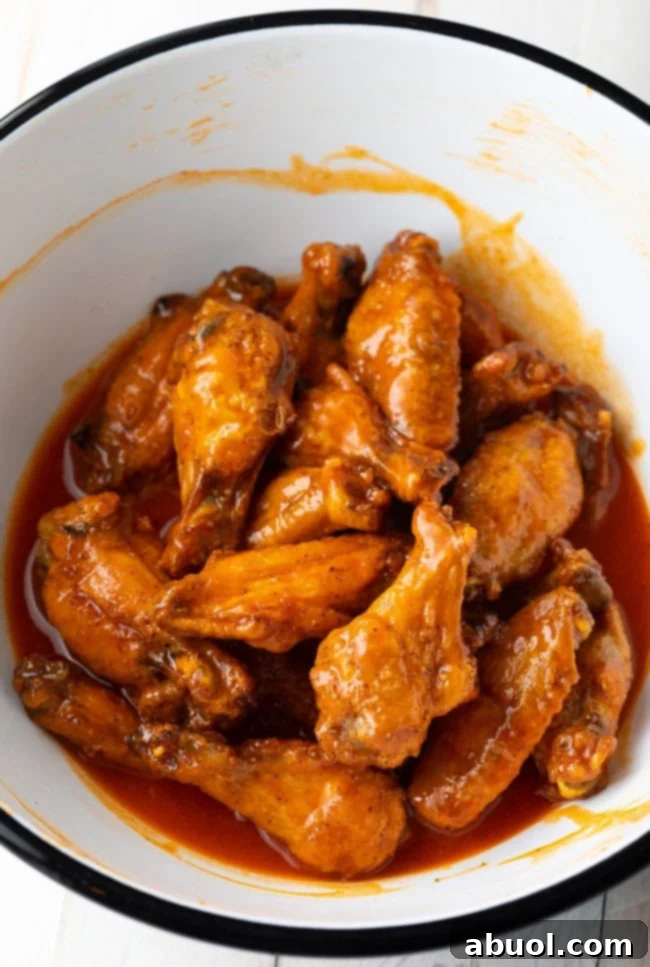 A large white mixing bowl filled with perfectly baked chicken wings, being vigorously tossed to ensure every piece is thoroughly coated in a vibrant, glossy buffalo sauce.