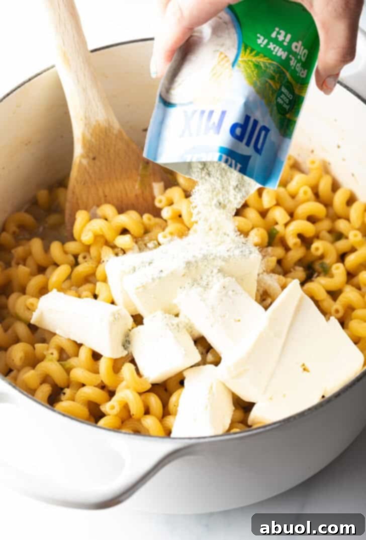 Large pieces of cream cheese and ranch dressing mix being added to a pot with simmering pasta.