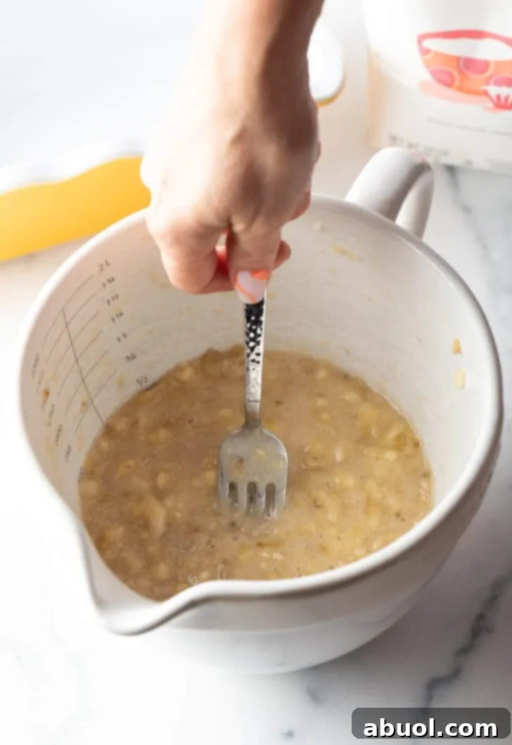 A hand expertly mashing ripe bananas in a white mixing bowl using a metal fork.