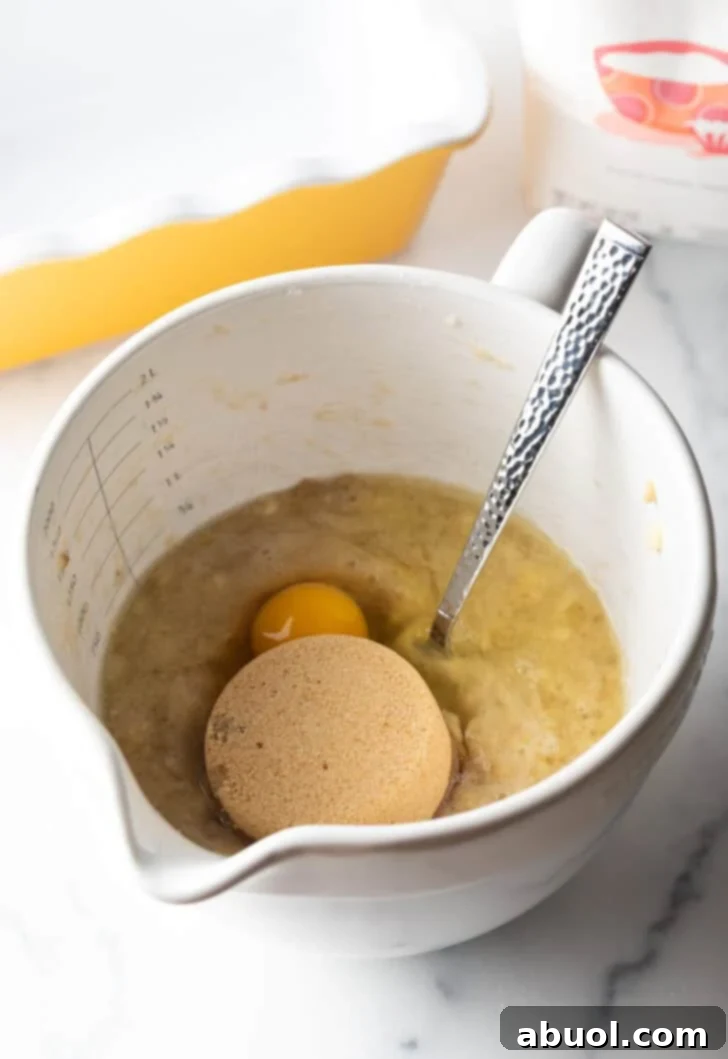 A scoop of rich brown sugar and two raw eggs being added to a large white mixing bowl filled with mashed bananas, ready to be combined.