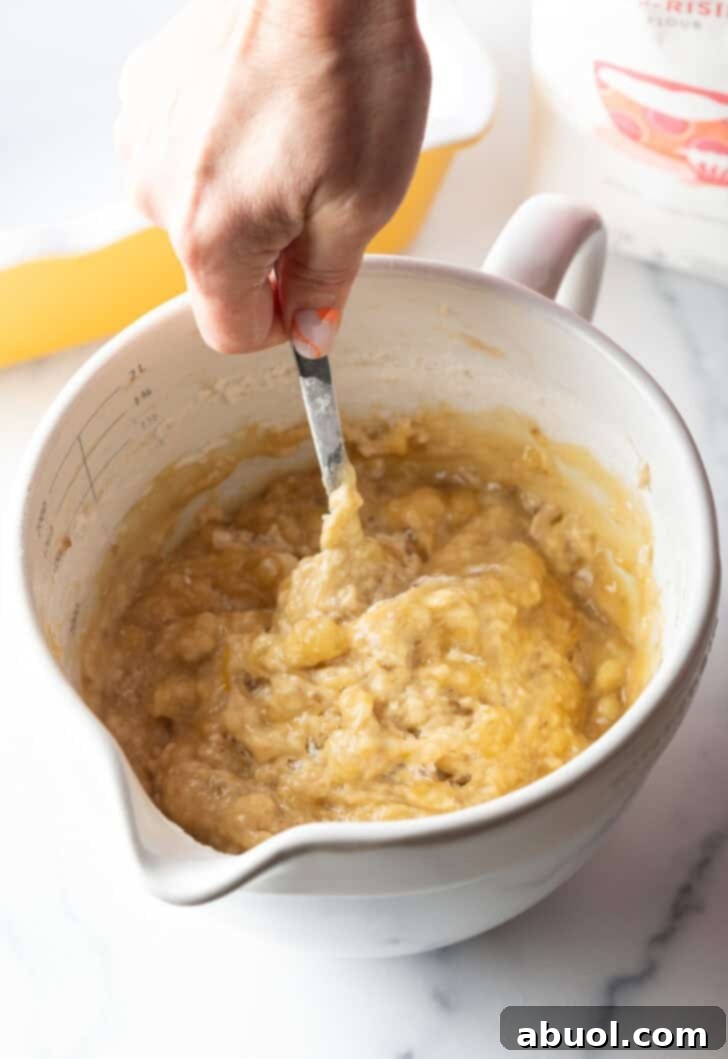 A hand gently stirring the banana bread batter in a large white mixing bowl with a metal spoon, ensuring all ingredients are just combined.