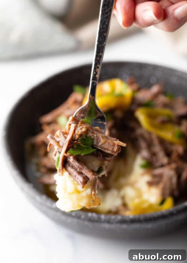A hand gently holds a fork, lifting a succulent piece of shredded pot roast with a pepperoncini towards the camera, showcasing its tender texture.