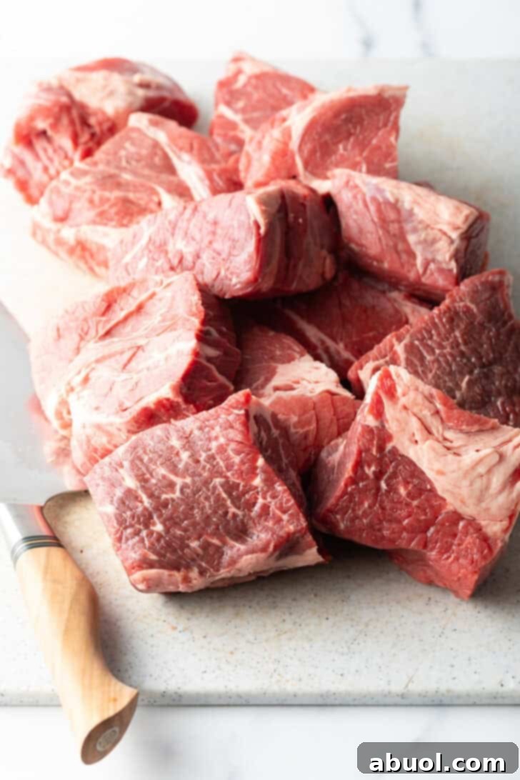 Large chunks of raw beef chuck roast resting on a wooden cutting board, alongside a sharp chef's knife, prepped for cooking.