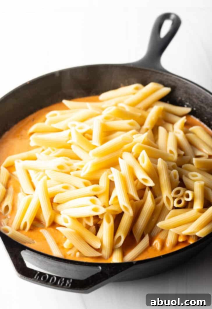 Al dente penne noodles being added to the luxurious cream sauce in a cast iron skillet, ready to be coated.