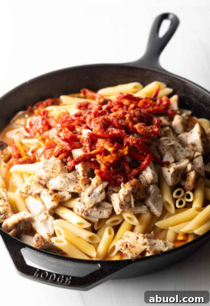 Chopped cooked chicken, sun-dried tomatoes, and fresh basil leaves being combined with penne pasta and cream sauce in a cast iron skillet.