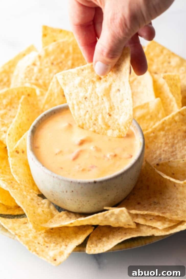 Hand dipping a tortilla chip into a pristine white bowl of golden Velveeta cheese dip, surrounded by a generous scattering of crunchy tortilla chips.