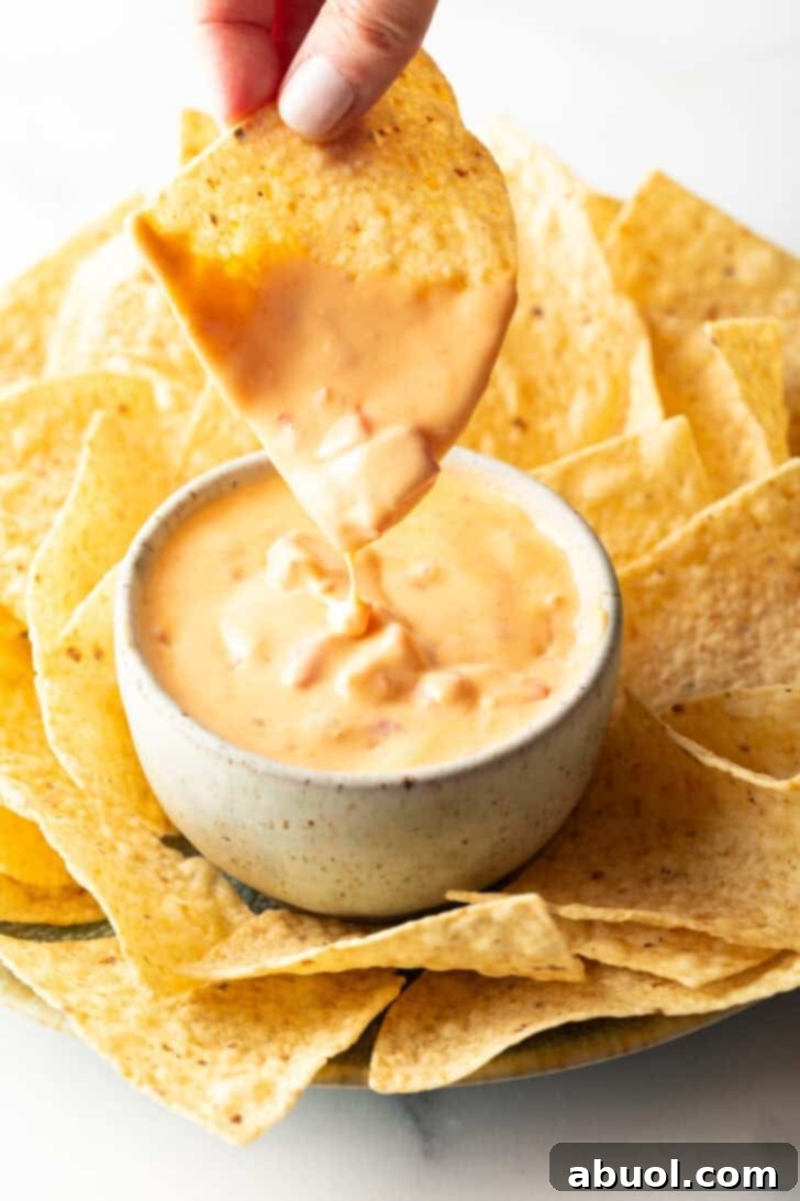 Hand dipping a chip into a white bowl of velveeta cheese dip, showing the rich, melted texture, surrounded by a scattering of golden tortilla chips.