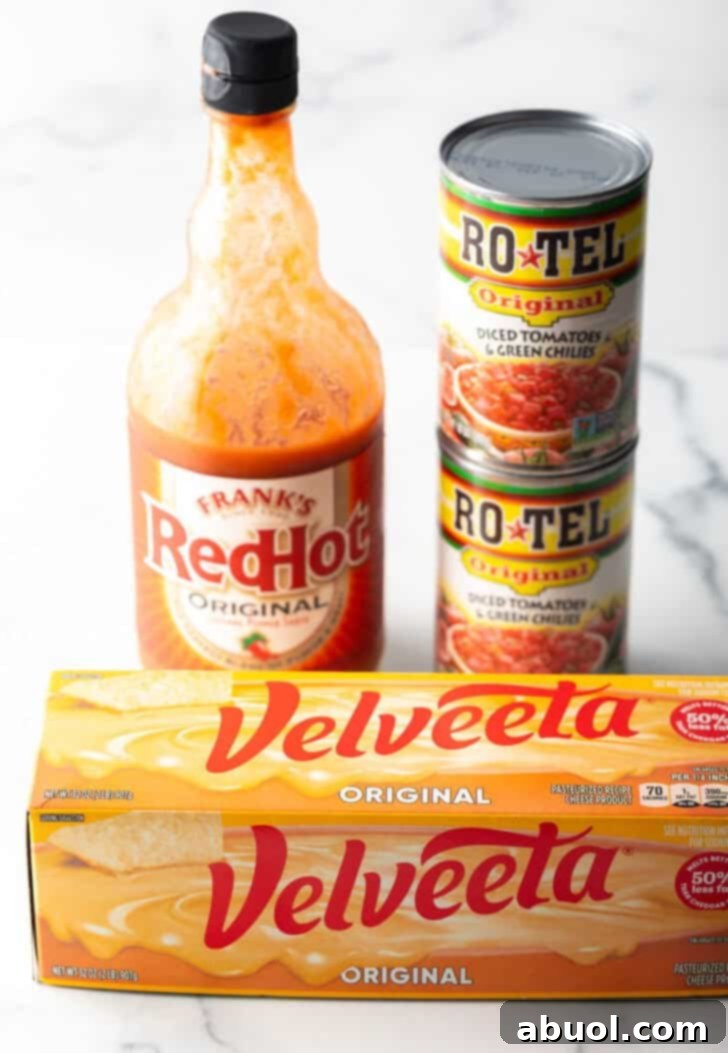 A block of Velveeta cheese, a bottle of Frank's RedHot sauce, and two cans of Rotel diced tomatoes and green chilies stacked on top of each other, showcasing the main ingredients.