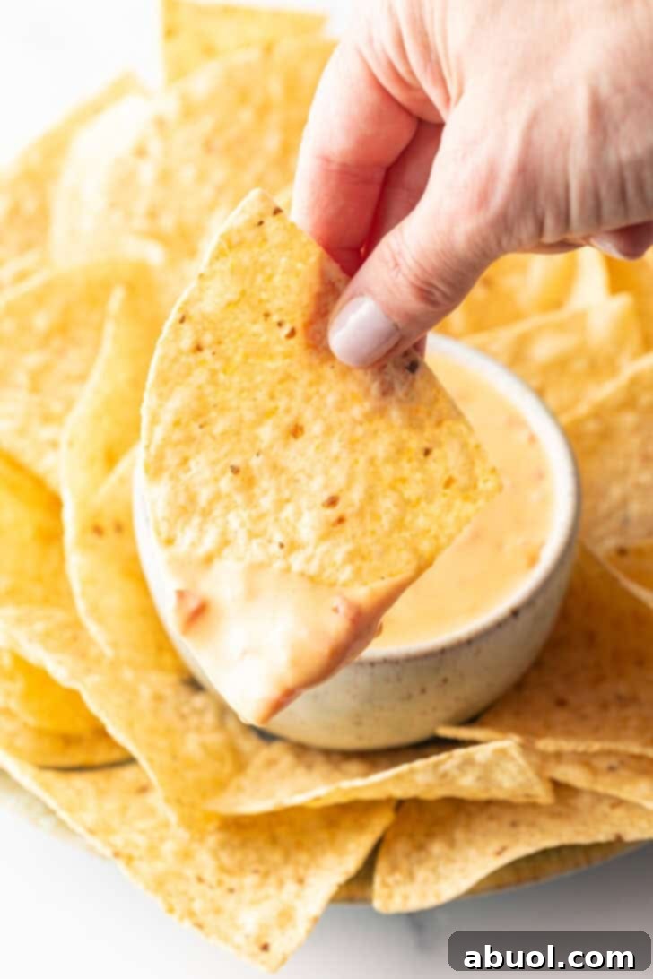 A hand holding a tortilla chip, dripping with golden Velveeta cheese dip, extended towards the camera, inviting a taste.