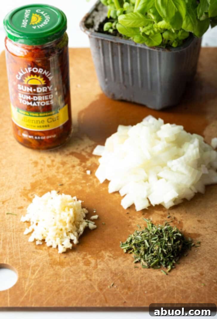 Tuscan Cream Chicken Delights 5 Recipe ingredients neatly arranged on a cutting board, including a jar of sun-dried tomatoes, diced onion, minced garlic, a bundle of fresh chopped herbs, and a box of baby spinach leaves, all ready for cooking.