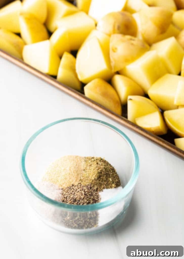 Raw pieces of cubed Yukon potatoes are spread evenly on a baking sheet, with a small glass dish to the side holding a vibrant mix of assorted spices.