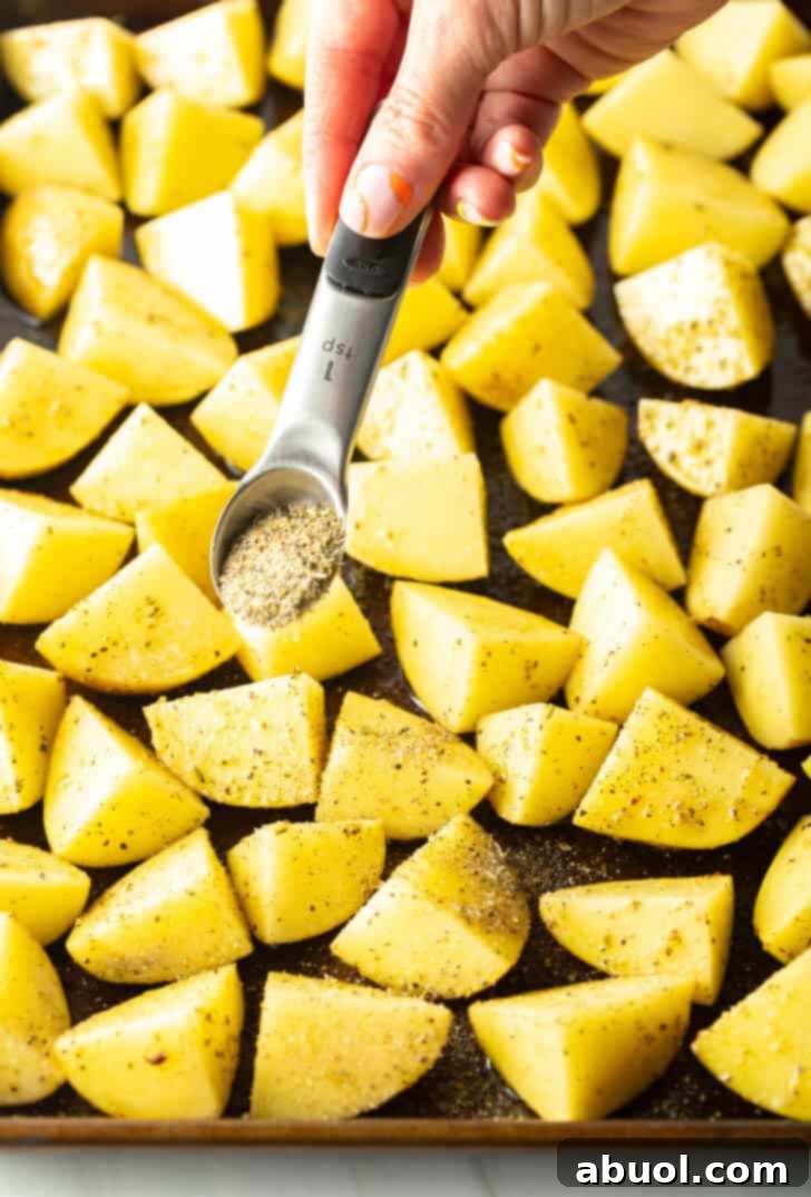 A hand gently sprinkles a seasoning blend from a small teaspoon over a generous scattering of potato chunks on a large baking sheet, ensuring even coverage.