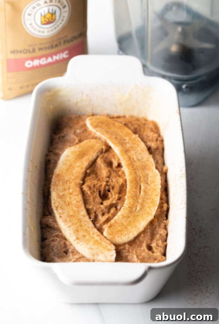 Banana bread batter in a white loaf pan, beautifully topped with two banana halves sliced lengthwise for an attractive finish.