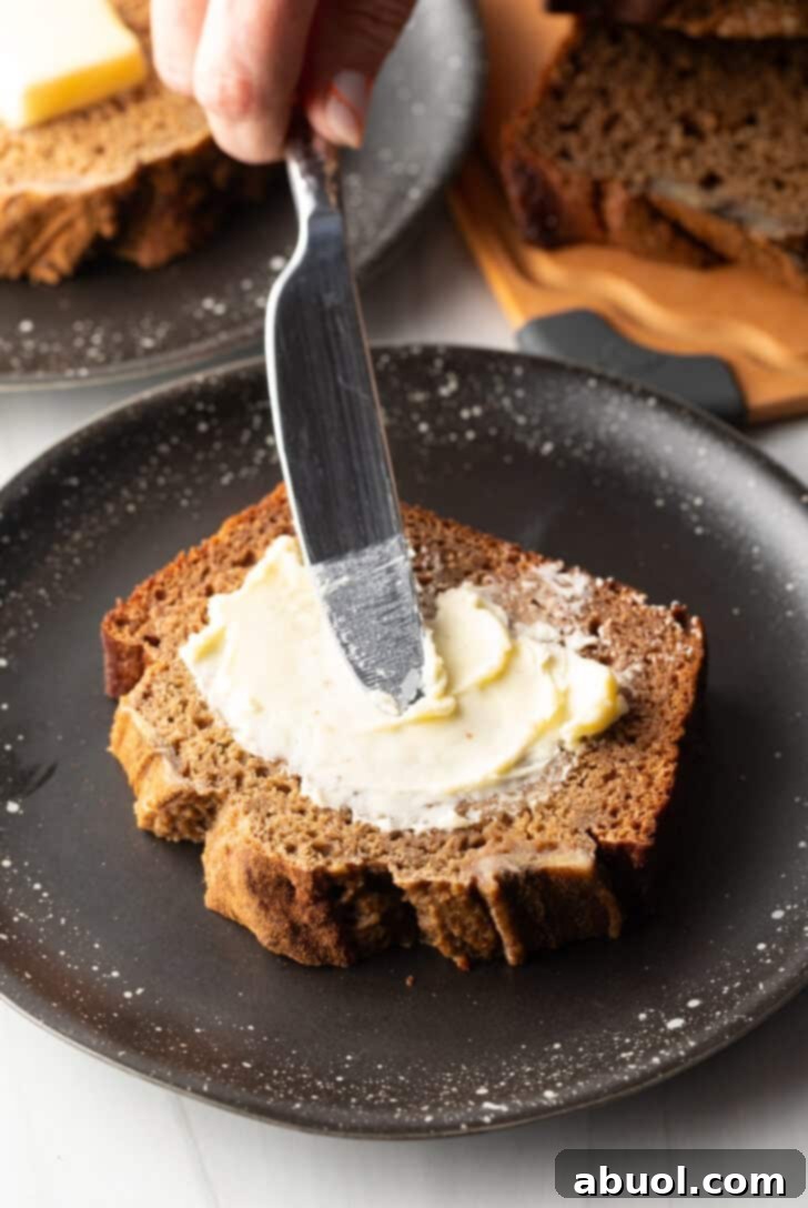 A hand using a knife to gently smear butter onto a thick, warm slice of healthy banana bread, artfully placed on a black plate.