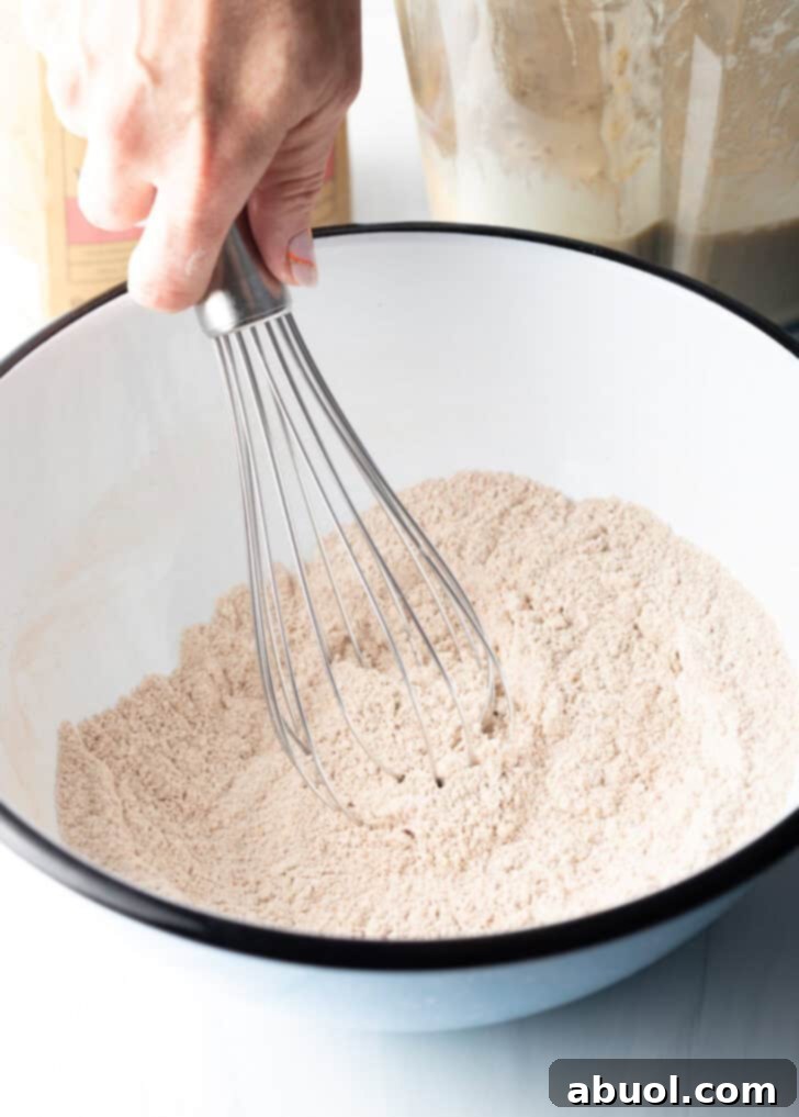 Whisking dry ingredients, including whole wheat flour, baking soda, cinnamon, and salt, in a white mixing bowl.