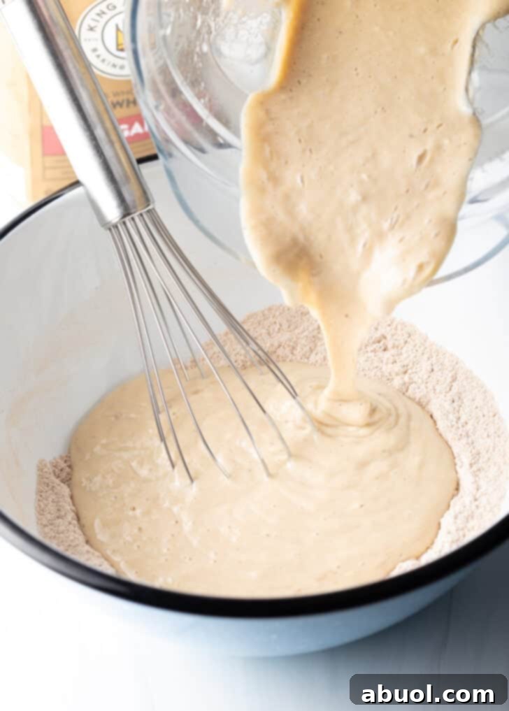 Pouring the blended wet ingredients from a glass bowl into the dry ingredients in a large white mixing bowl.