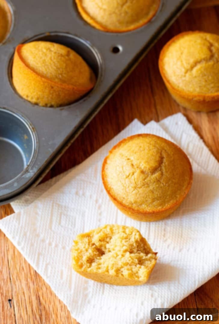 Two honey cornbread muffins on a white napkin, next to a muffin baking tin with more muffins. One muffin has been split in half to show the inside.