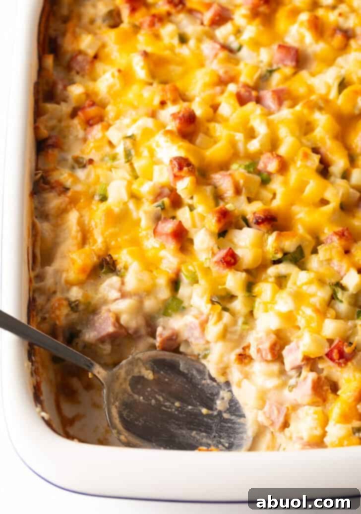 Close-up, top-down view of a white casserole dish filled with golden brown cheesy ham and potato casserole, with a serving spoon removing a portion.