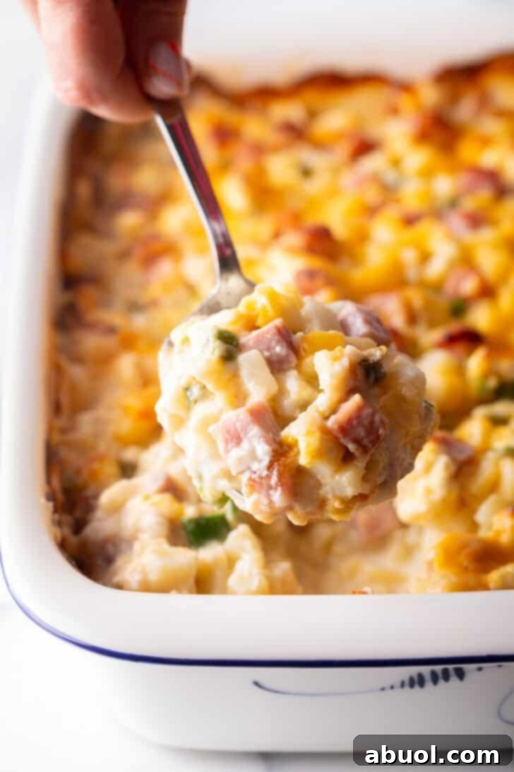 A hand holding a metal spoon, presenting a spoonful of cheesy ham and potato casserole directly to the camera.