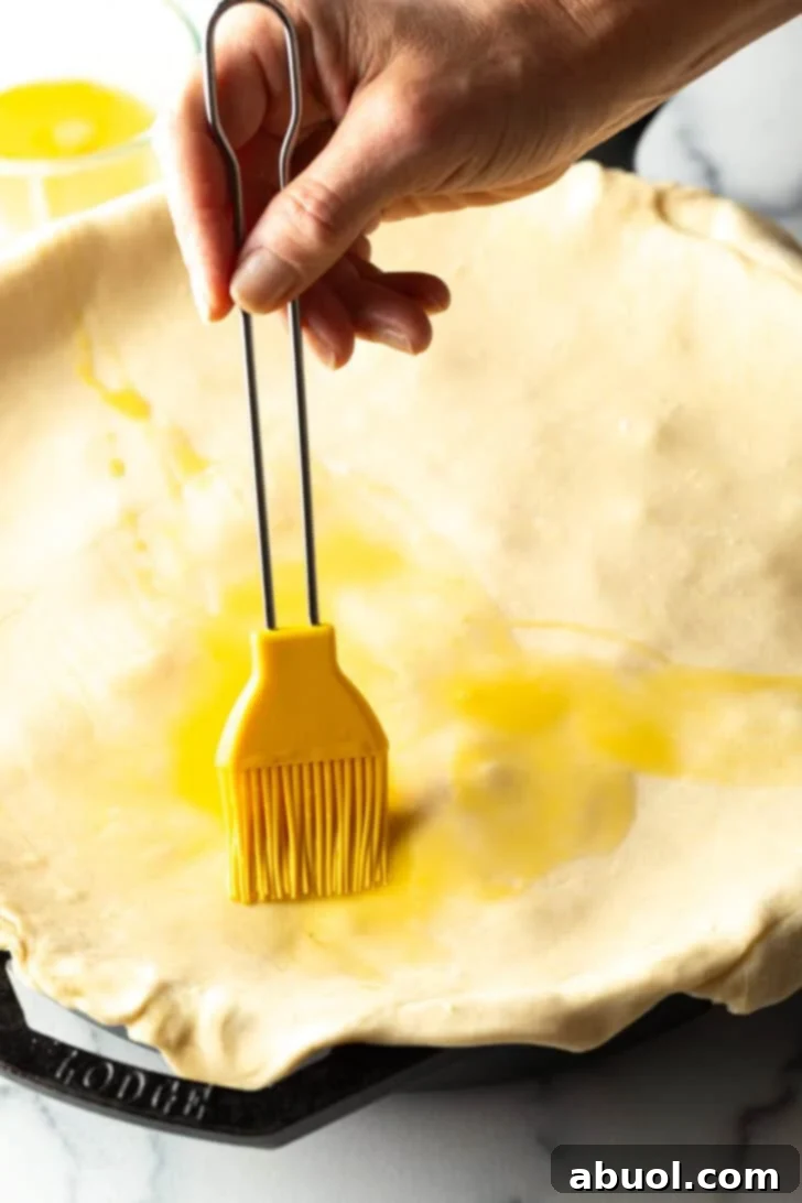 Hand using a yellow pastry brush to coat the top of puff pastry with melted butter.