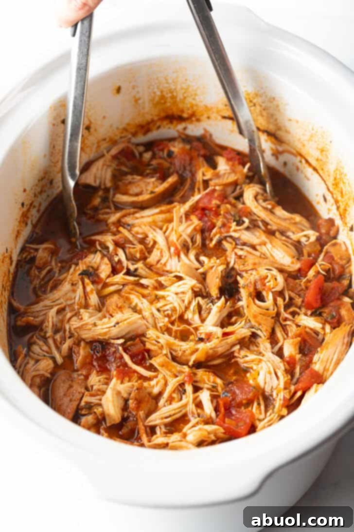 Shredded Mexican chicken in a white crockpot with tongs, ready to serve.