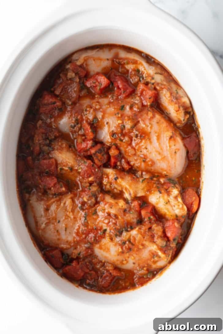 Several whole chicken breasts covered in red sauce in a white crockpot, ready for cooking.