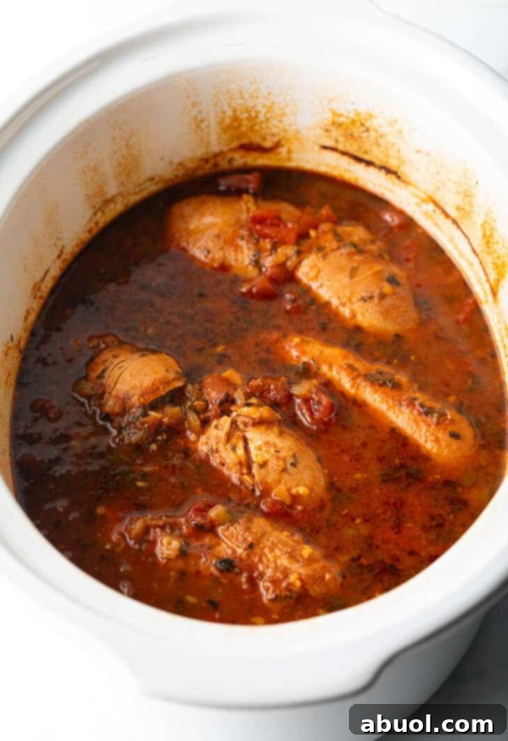 Several whole chicken breasts covered in red sauce in a white crockpot, midway through cooking.
