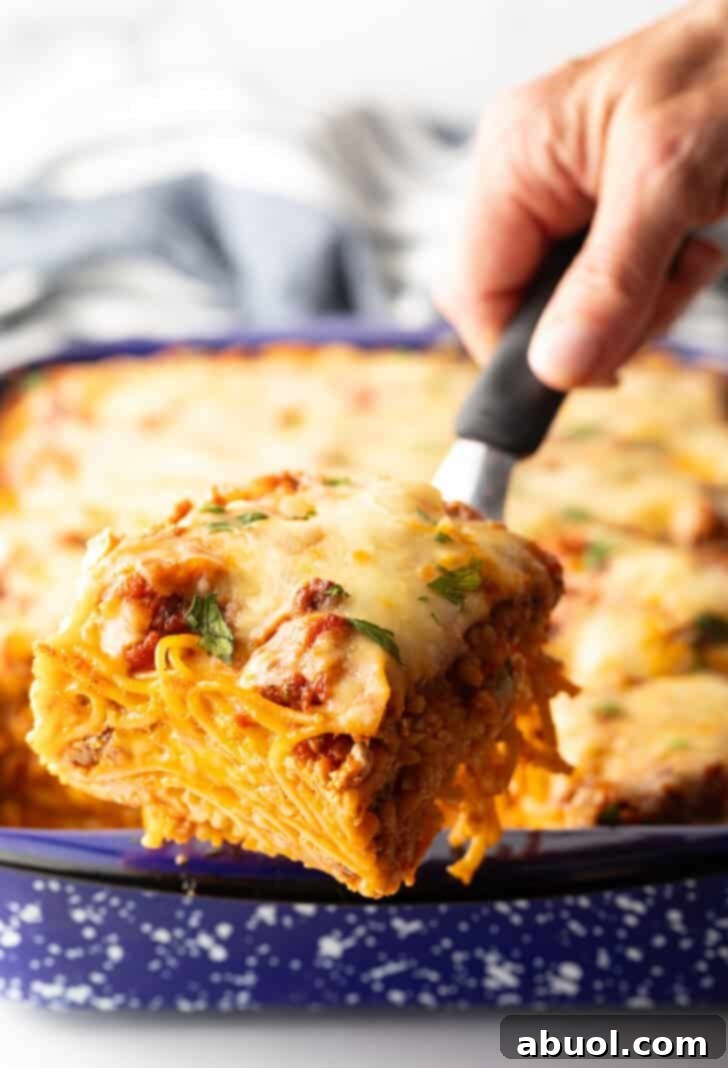 Hand with a spatula holding a serving of baked spaghetti to camera, showcasing the cheesy layers.