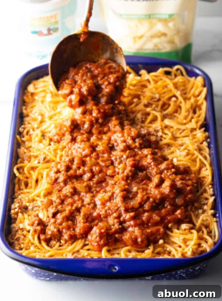 A ladle pouring the remaining rich meat sauce over the prepared spaghetti mixture in a blue casserole dish, creating a top layer.