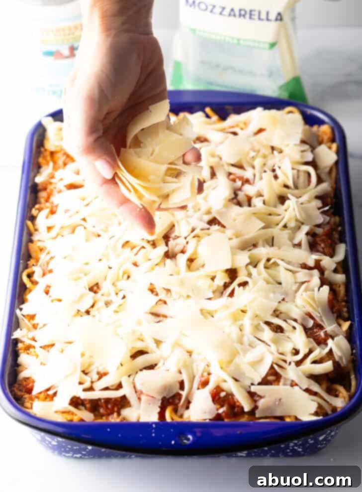 A hand adding a generous sprinkle of shredded parmesan cheese on top of the spaghetti casserole, just before baking.