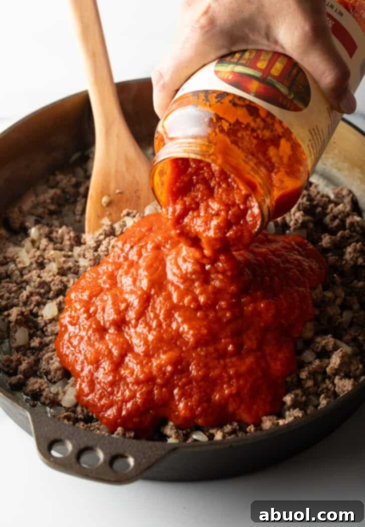 Adding jarred marinara sauce to a skillet with browned ground beef, onions, and garlic, creating the base for the meat sauce.