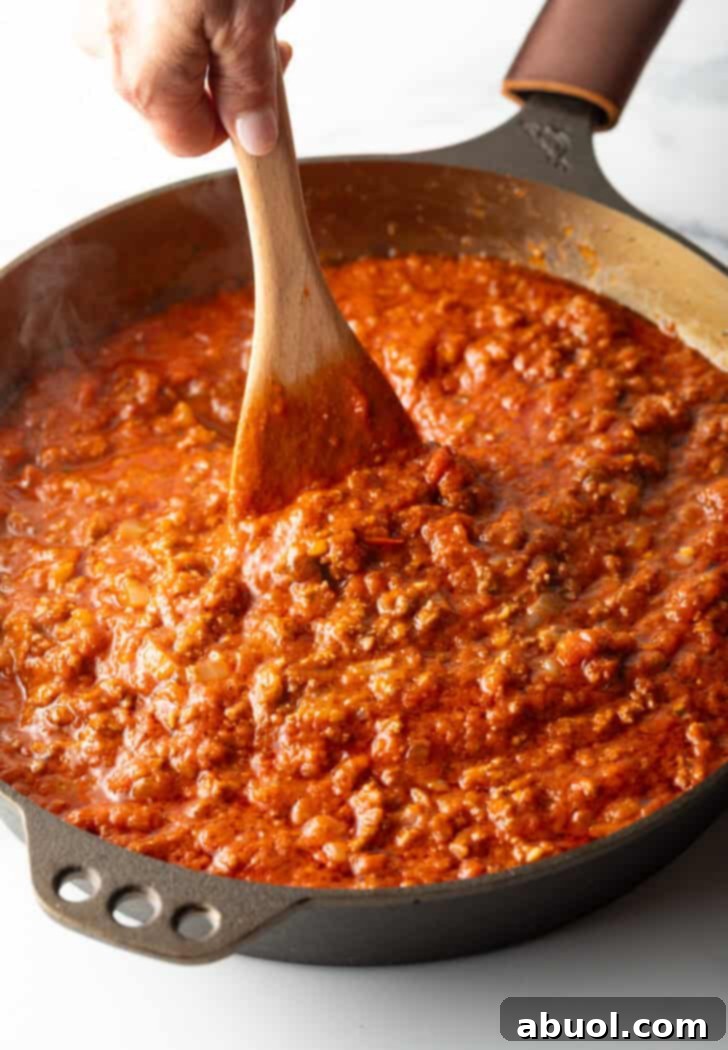 Stirring the rich meat pasta sauce with a wooden spatula in the skillet, ensuring all ingredients are well combined and simmering.