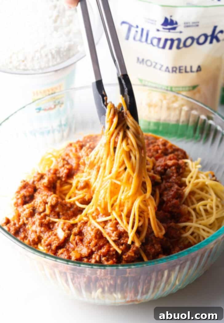 Tongs tossing cooked spaghetti together with half of the savory meat sauce in a large glass bowl, ensuring even coating.