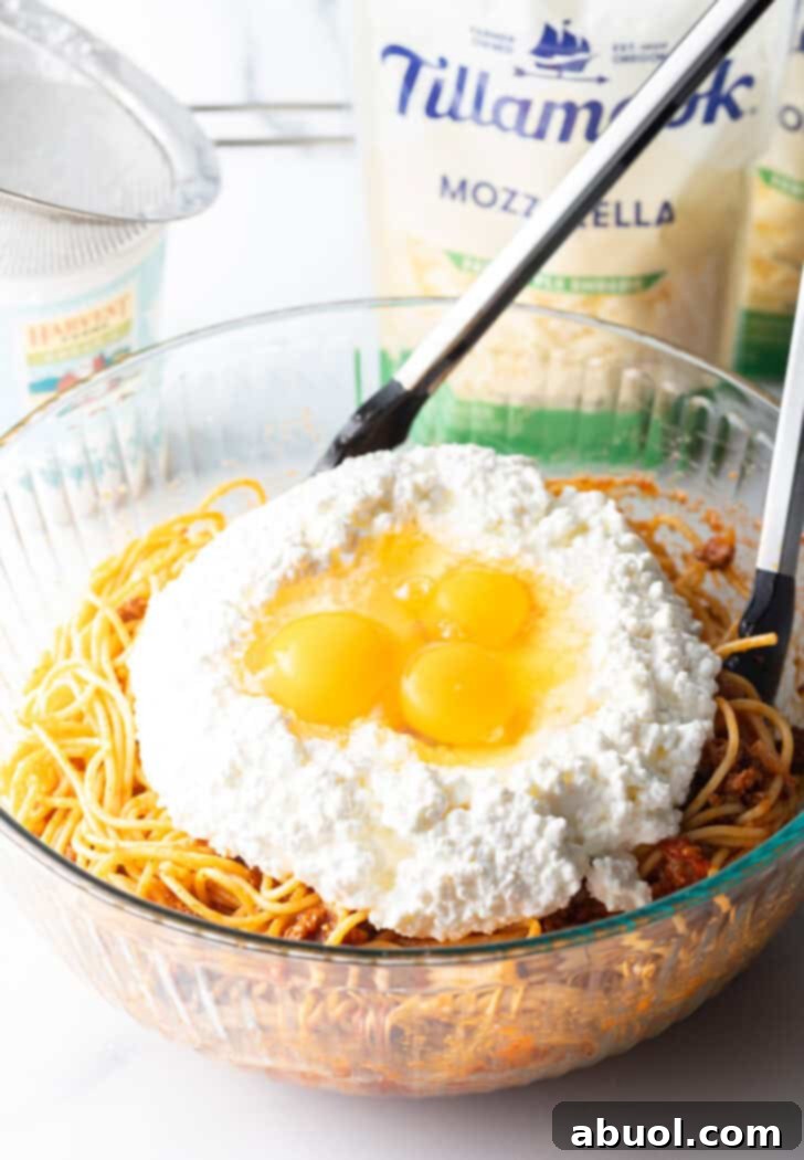 A large glass bowl holding cooked spaghetti, with creamy cottage cheese and three cracked eggs ready to be mixed in.
