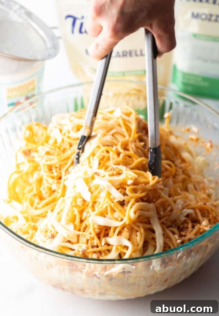 Using tongs to thoroughly toss cooked spaghetti in a glass bowl with the cottage cheese, eggs, and mozzarella mixture.