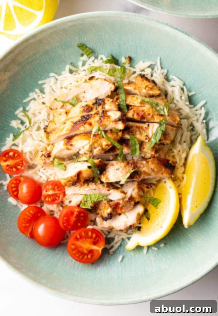 Shallow blue bowl with white rice topped with slices of marinated Greek chicken, halved grape tomatoes, and two lemon wedges.
