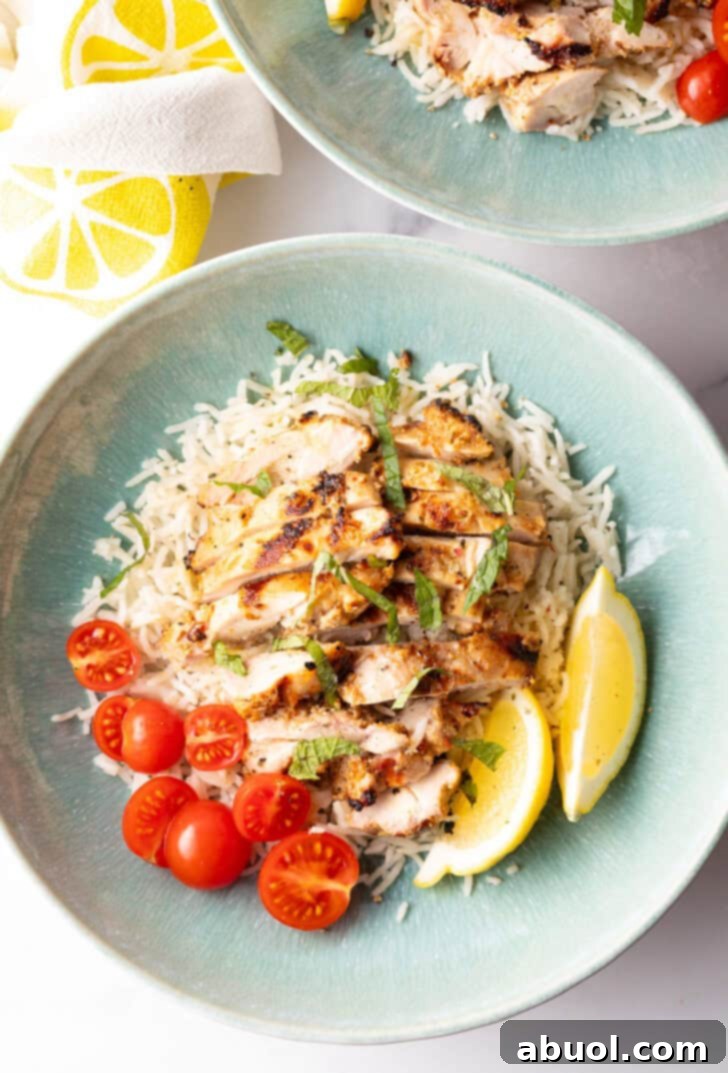 Shallow blue bowl with white rice topped with slices of marinated Greek chicken, halved grape tomatoes, and two lemon wedges, ready to eat.