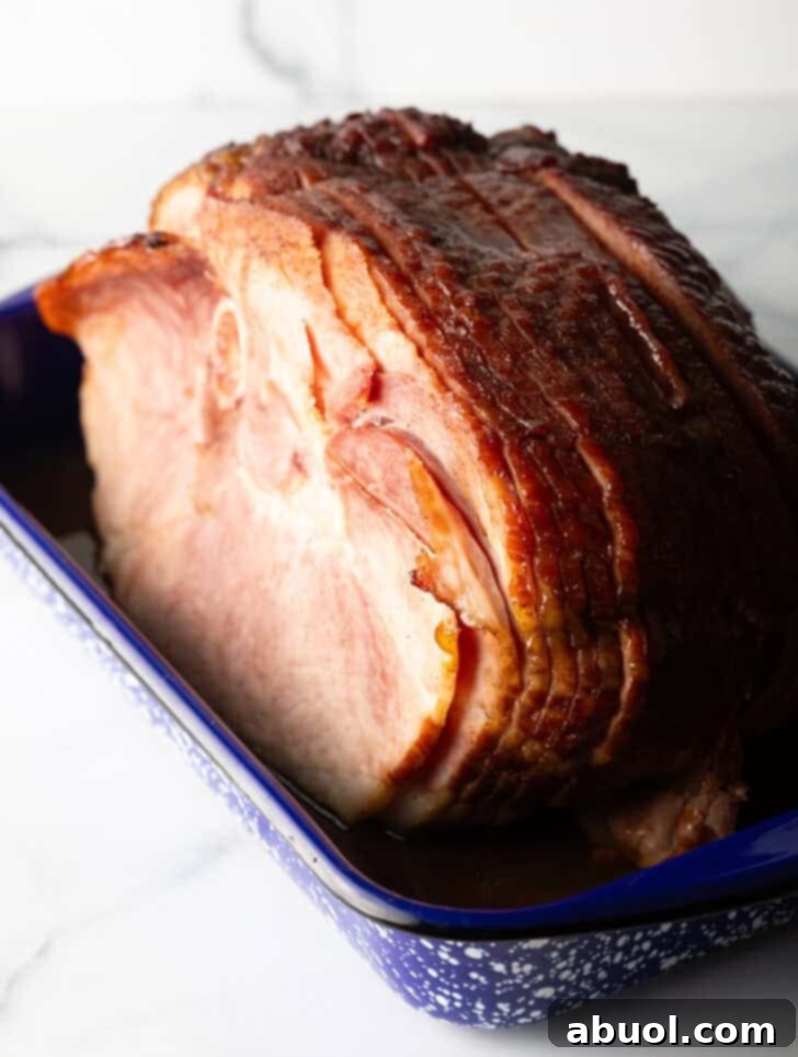 A large cooked spiral ham, beautifully glazed and ready for carving, in a blue and white speckled roasting pan, garnished with herbs.