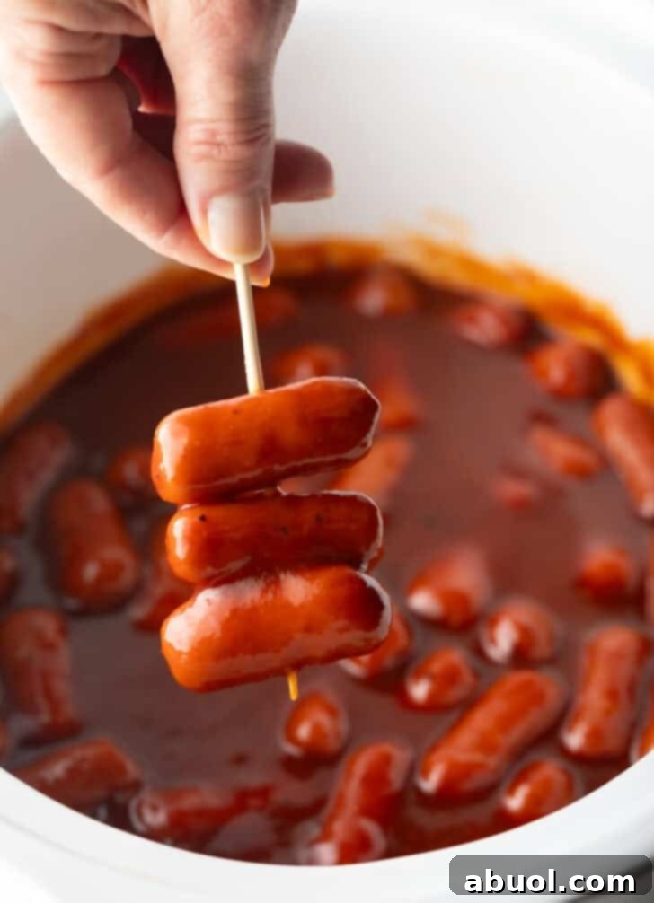 Hand holding a skewer with 3 little smokies, over a crock pot with cooked mini sausages in bbq sauce.