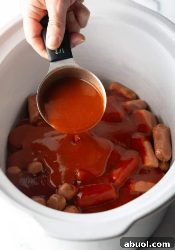 Hand pouring hot sauce from a measuring cup into a slow cooker with smokies.