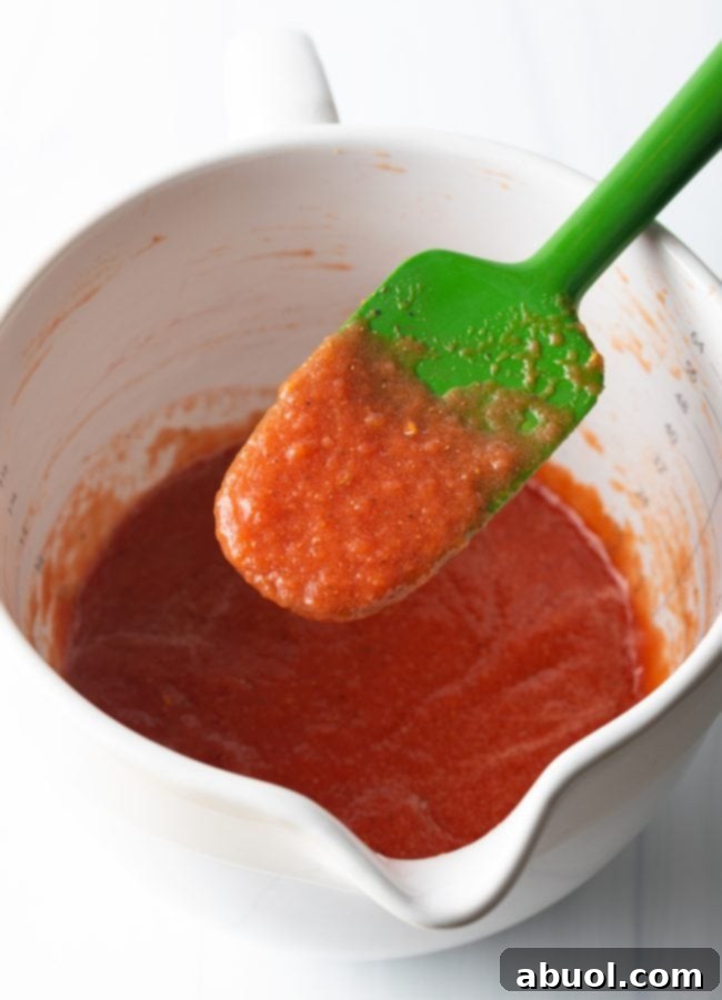 Crafting Perfect Neapolitan Pizza Dough 11 White mixing pitcher filled with tomato sauce. A green spatula is covered in the sauce and held to camera.