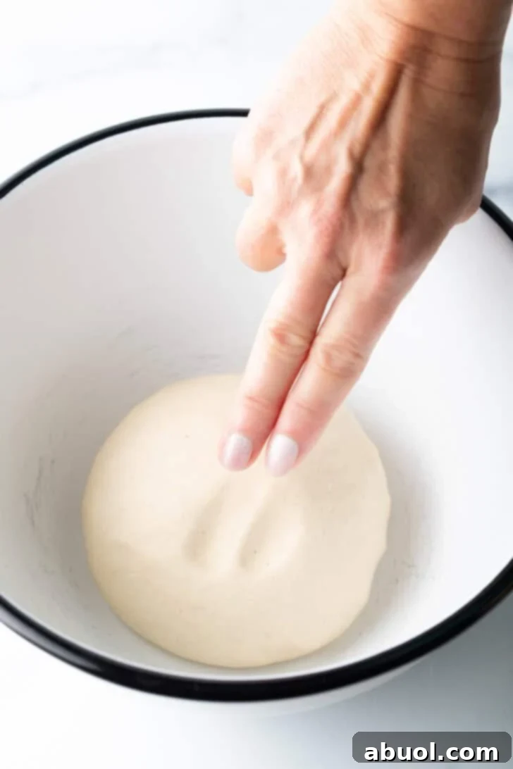 Crafting Perfect Neapolitan Pizza Dough 6 Hand using two fingers to poke a round of pizza dough in a large white mixing bowl.